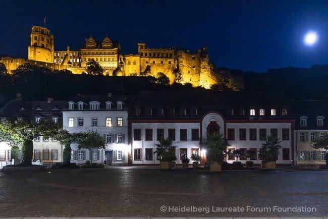 White row houses in Germany on the street level. A castle lit up at night is behind and on a hill.  