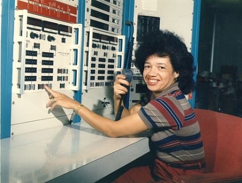 A photo of a smiling woman with light-brown skin and dark hair in front of a dated computer. 