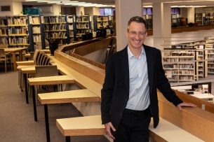 Centenary University psychology professor Tal Ben-Shahar, a light-skinned man with short hair wearing glasses, stands in front of shelves of books.