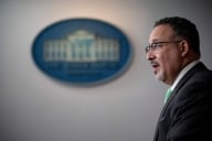 Education Secretary Miguel Cardona stands at a podium. A blurred-out image of the White House in the background.