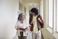 Front view of curly-haired man with backpack side by side with mature female professor and conversing as they approach in hallway.