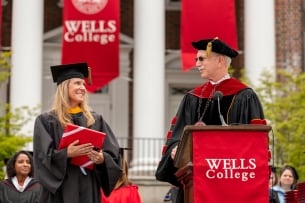 Wells College president Jonathan Gibralter, a light-skinned man with gray hair and a mustache, wearing academic regalia and sunglasses, on graduation day in front of red Wells College signage.