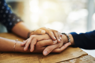 A pair of hands with darker skin resting on a table hold a hand with lighter skin in a comforting manner