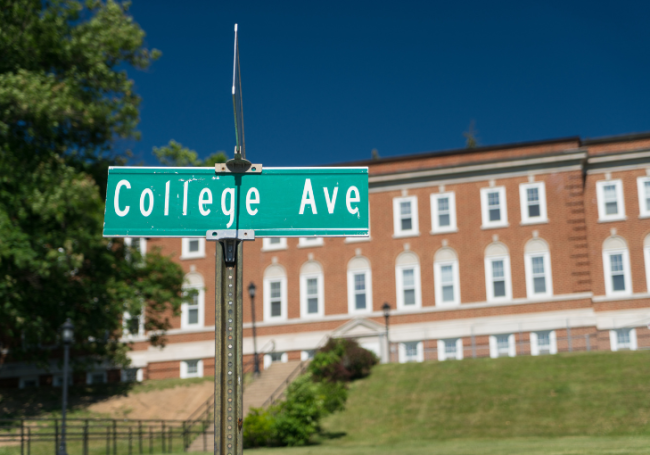 A close-up of a green road sign that says "College Ave" with a brick university building in the background.